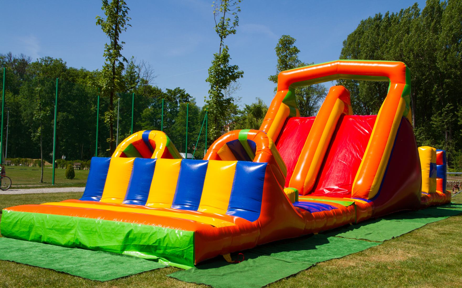 Long inflatable obstacle course with a tall slide, staked on a ground tarp in a park